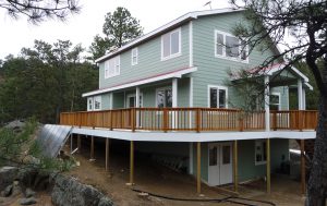Shown here is a custom off-grid home with second story wrap-around wood deck and solar panels on the ground.